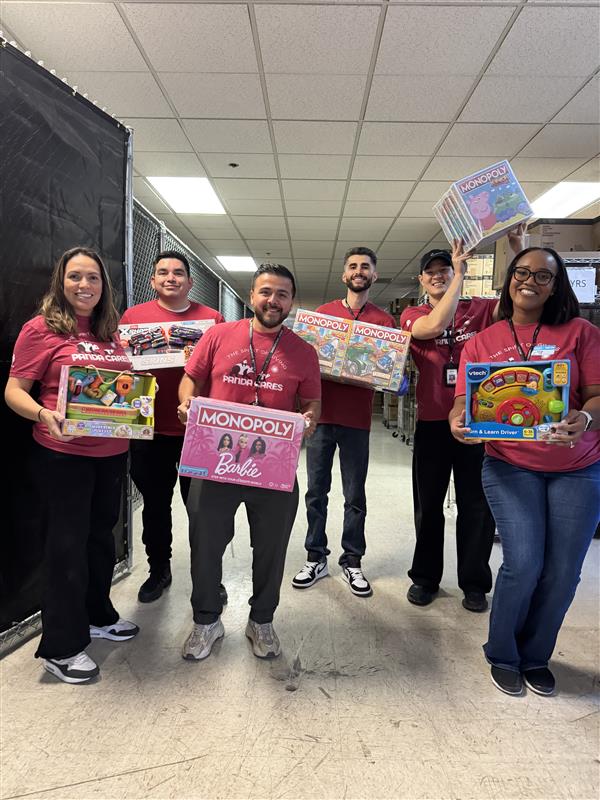 Group of Panda Cares volunteers in matching red shirts holding donated toys and board games, including Monopoly and children’s learning toys, during a volunteer gift sorting event